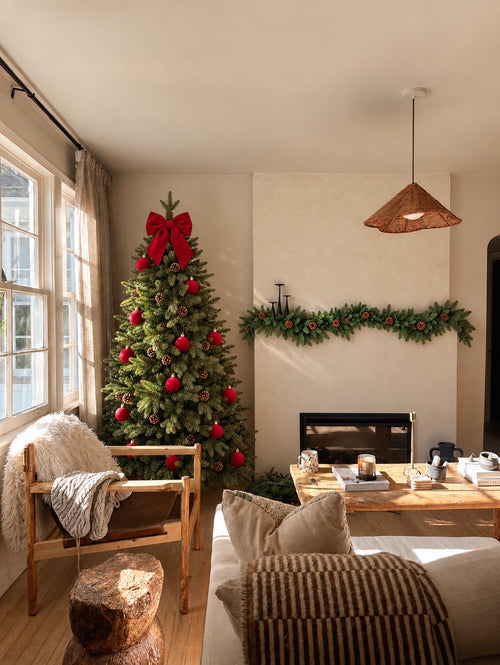 Abeto artificial balsámico decorado con lazo y bolas rojas en un salón, con guirnalda balsámica sobre la chimenea · BOSSVI