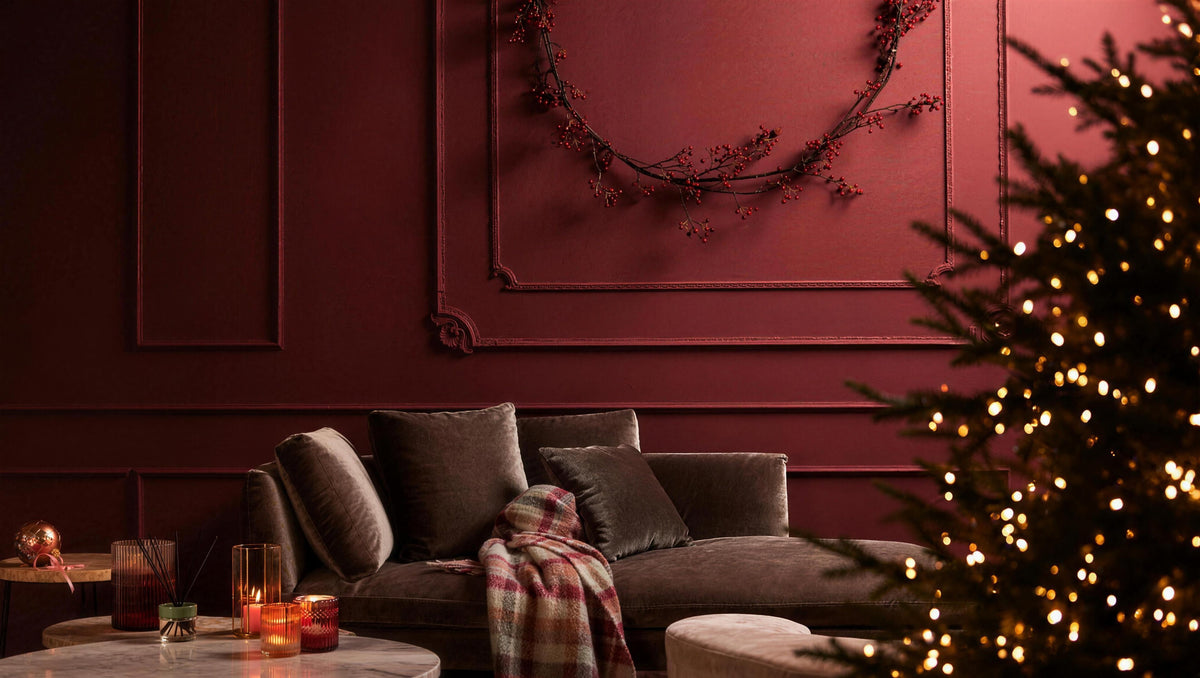 Salón con pared burdeos con molduras, sofá de terciopelo, corona de bayas rojas y un abeto de Navidad con luces
